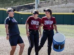 Two softball players laugh on the field with Coach Shena Hollar